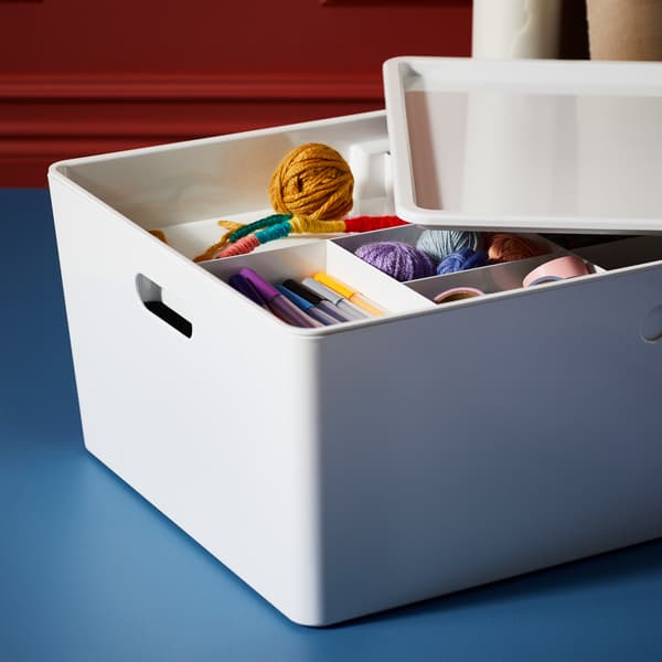 Una caja KUGGIS blanca que contiene una selección de plumas de colores, cinta adhesiva rosa y varios ovillos de hilo de colores vivos.