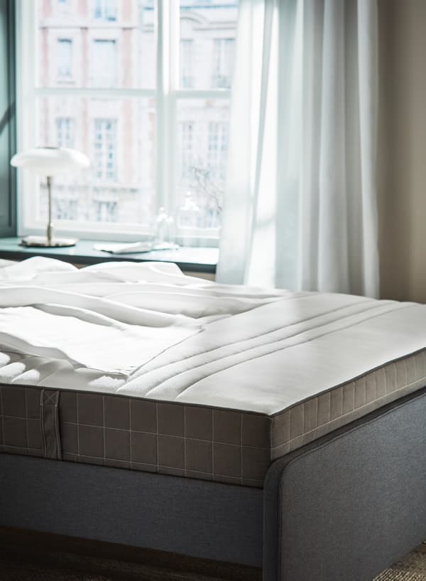 Una cama al descubierto con unas sábanas blancas encima. Al fondo, vemos una ventana que da a la calle y se puede ver la fachada del edificio de enfrente.