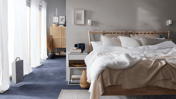 Una cama BJÖRKSNÄS de abedul con ropa de cama blanca y beige junto a una mesita de noche blanca, una lámpara de mesa de bambú y un purificador de aire.