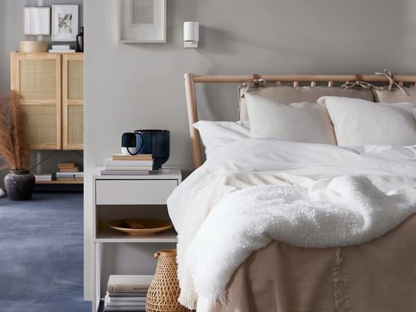Una cama BJÖRKSNÄS de abedul con ropa de cama blanca y beige junto a una mesita de noche blanca, una lámpara de mesa de bambú y un purificador de aire.
