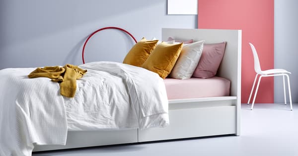 Una cama blanca MALM con cajones de almacenamiento, ropa de cama blanca y almohadas en amarillo, rosa y blanco.