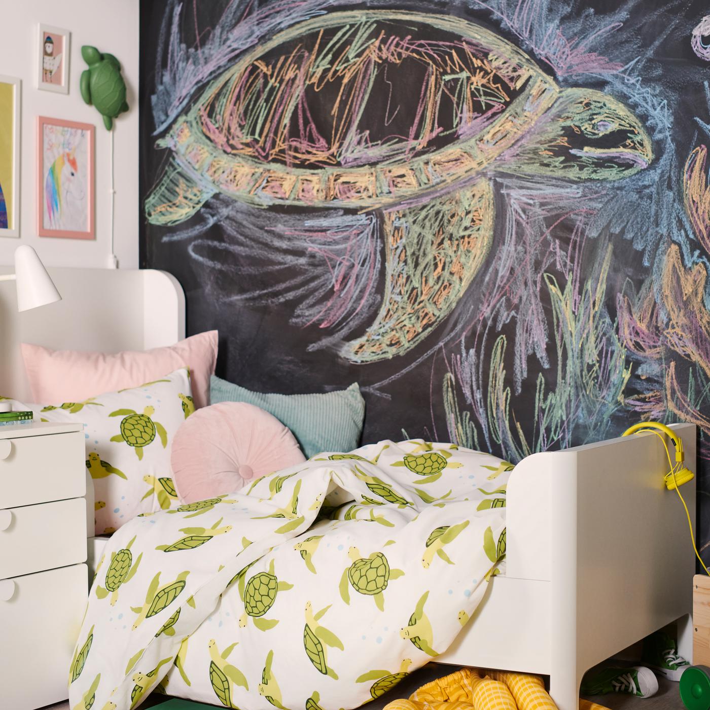 Una cama BUSUNGE con la funda nórdica BLÅVINGAD con un motivo de tortugas verdes y varios cojines, delante de una pared convertida en pizarra.