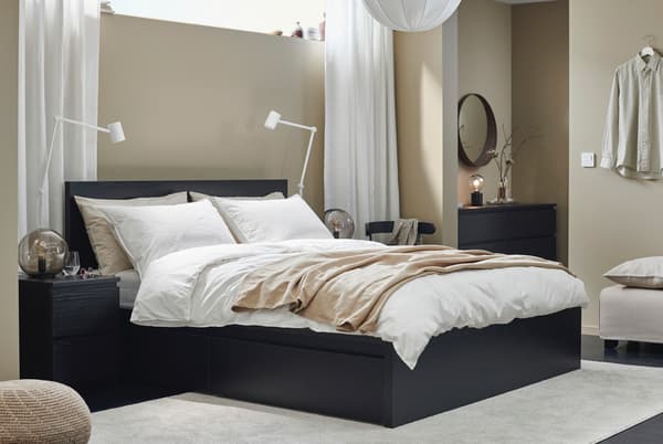 Una cama café oscuro con ropa de cama de color claro en una habitación con cortinas blancas y pared beige.