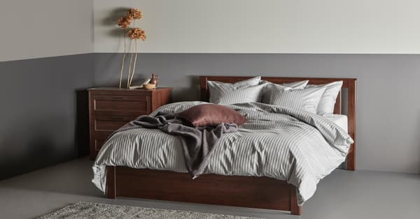 Una cama café SONGESAND con ropa de cama gris y una cómoda café de 3 cajones junto a ella.