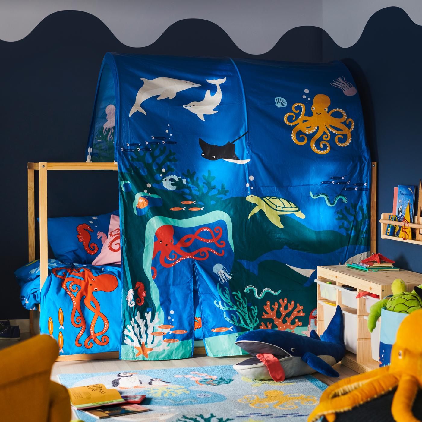 Una cama con un dosel KURA de animales acuáticos; delante hay una alfombra BLÅVINGAD con una ballena azul de peluche BLÅVINGAD.