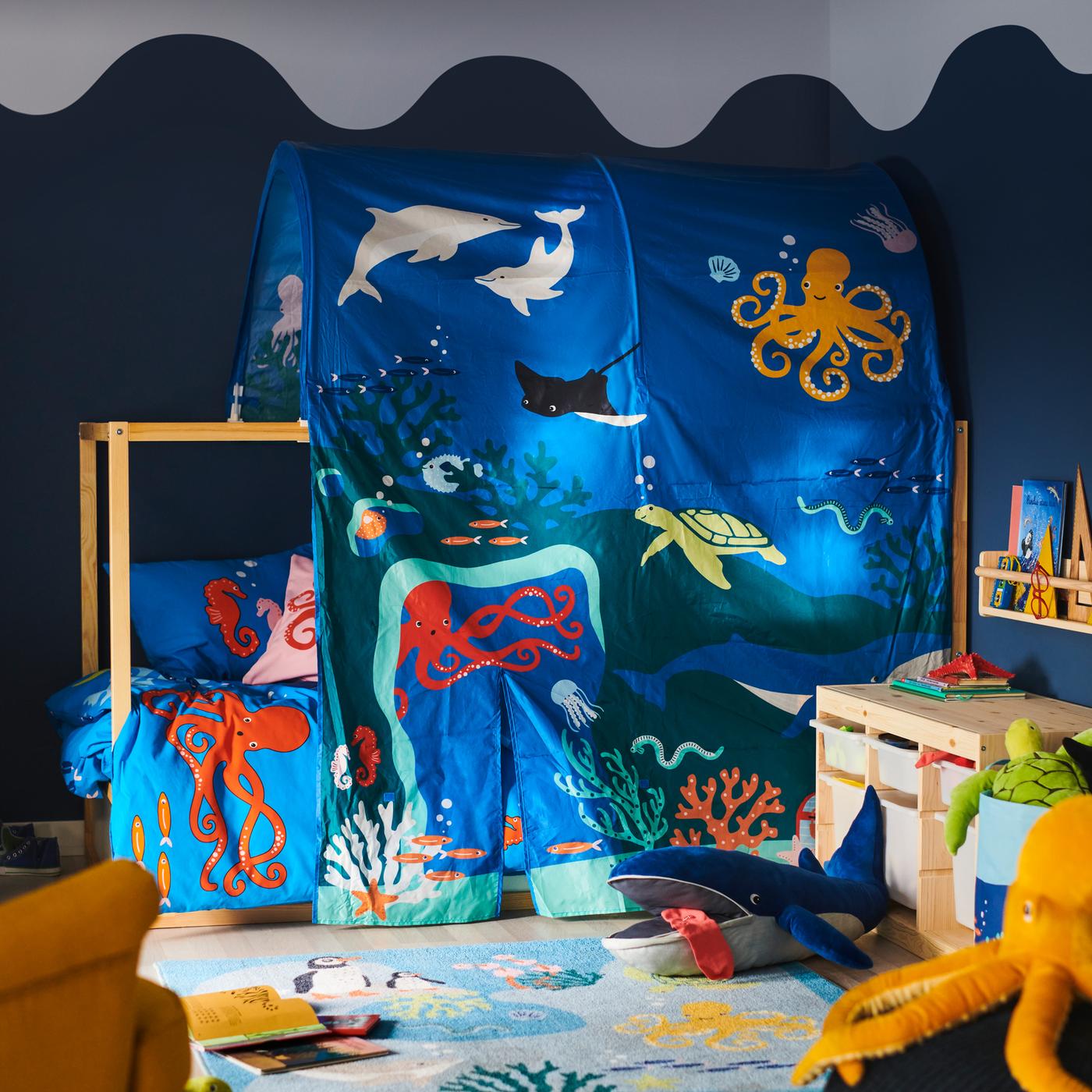 Una cama con un dosel KURA de animales acuáticos; delante hay una alfombra BLÅVINGAD con una ballena azul de peluche BLÅVINGAD.