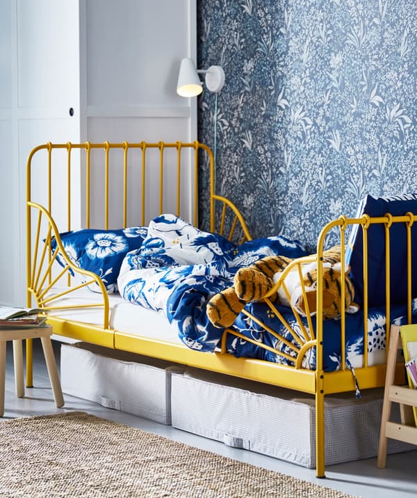 Una cama de niño con estructura amarilla y ropa de cama con motivos florales en azul, con cajas debajo y un aplique arriba.