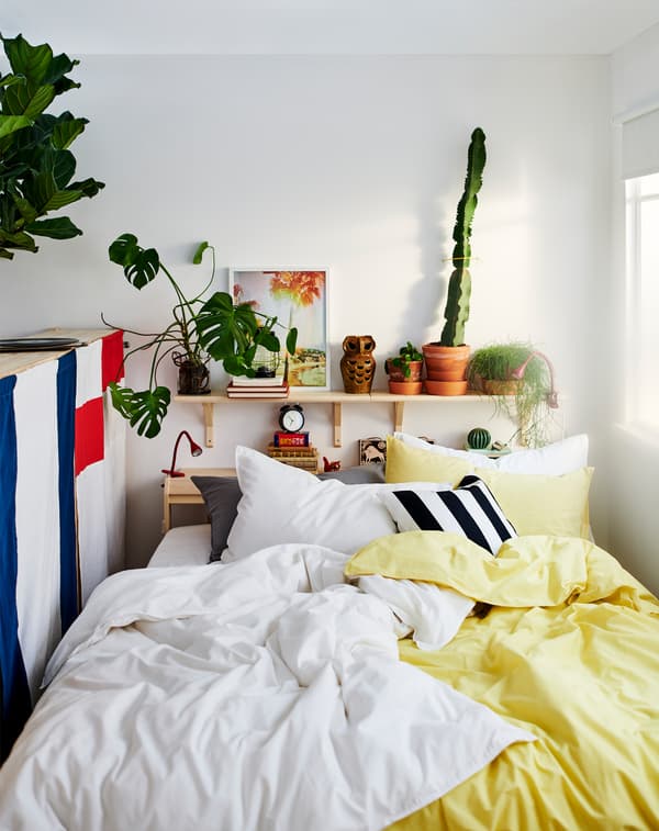 Una cama entre una ventana y la parte trasera de un módulo de almacenaje, con muchas almohadas y ropa de cama en amarillo, blanco, negro y gris.
