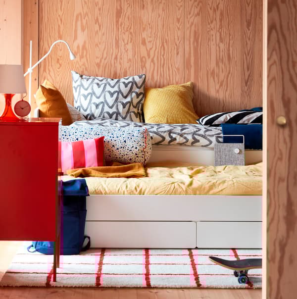 Una cama SLÄKT extensible en un dormitorio con paneles de madera vestida con un edredón amarillo y otro blanco con corazones grises.