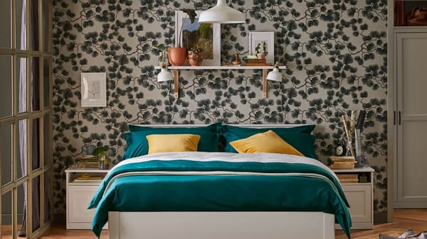 Una cama tamaño full con ropa de cama verde y cojines amarillos. Detrás de ella hay una estantería en la pared y una lámpara colgante encima.