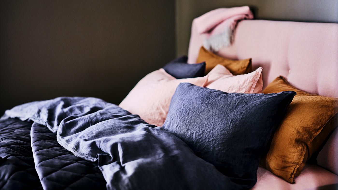 Una cama tapizada IDANÄS de color rosa pálido con almacenamiento, cubierta con un edredón gris oscuro y almohadas con sábanas de color rosa pálido.