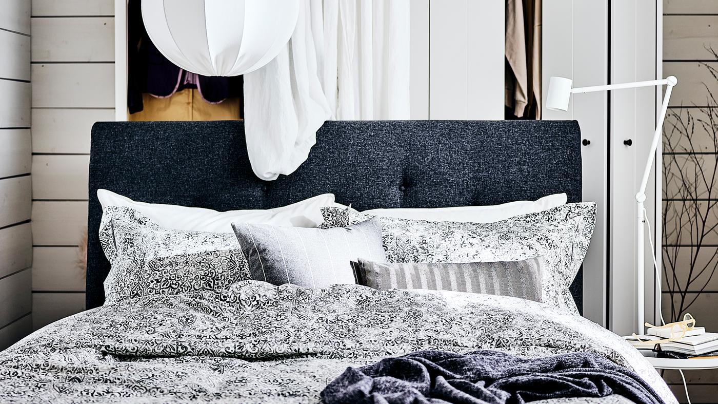 Una cama tapizada IDANÄS en gris oscuro con una cortina blanca, un armario blanco detrás y una lámpara de techo blanca encima.