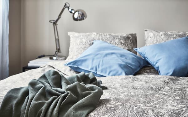 Una camera da letto con cuscini e un plaid. Accanto, un comodino con una lampada da lavoro nichelato FORSÅ.