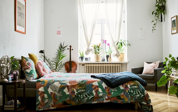 Una camera da letto con tante piante, tende sottili alla finestra, un letto con un piumino dalla fantasia tropicale e il pavimento in parquet.