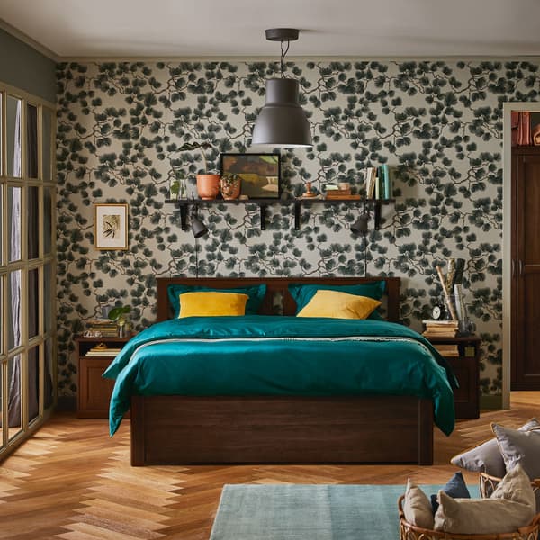 Una camera da letto con una parete con un motivo vegetale. Al centro un letto con due comodini con sopra dei libri. Attaccata alla parete una mensola e con sopra diversi oggetti. 