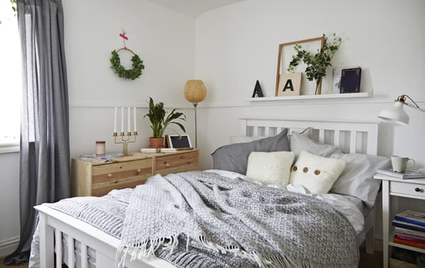 Una camera da letto di colore bianco e grigio con diversi cuscini. A lato, cassettiere in legno con sopra un vaso e un candeliere con candele. Accanto c'è una tenda grigia aperta.