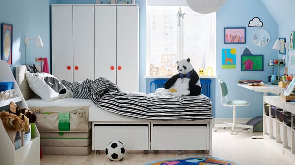 Una cameretta sui toni del bianco e con le pareti azzurrre. Al centro, c'è un letto con non ancora sistemato. A lato, una scrivania con mobli contenitori.