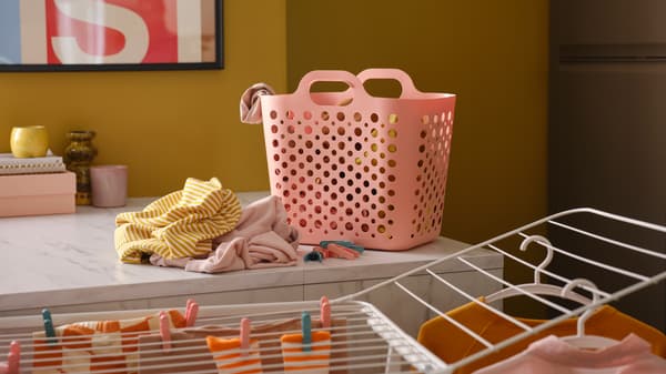Una canasta SLIBB para la ropa de color rosa y un montón de ropa sobre una cubierta de cocina, junto a un tendedero blanco en el que hay más ropa.