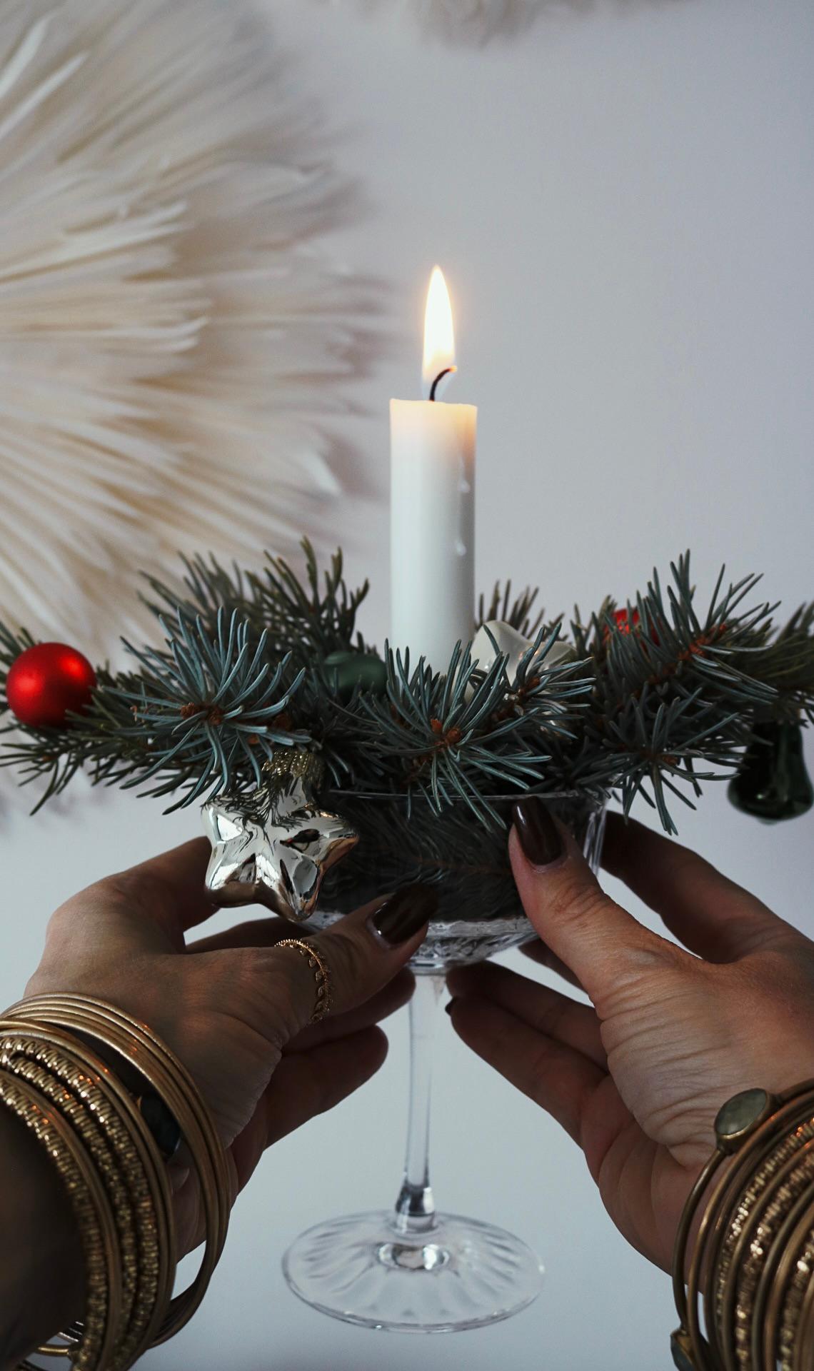 Una candela bianca in un portacandele di vetro è decorata con rami di pino e palline rosse. È tenuta da mani con braccialetti, che trasmettono un'atmosfera festosa ed elegante.