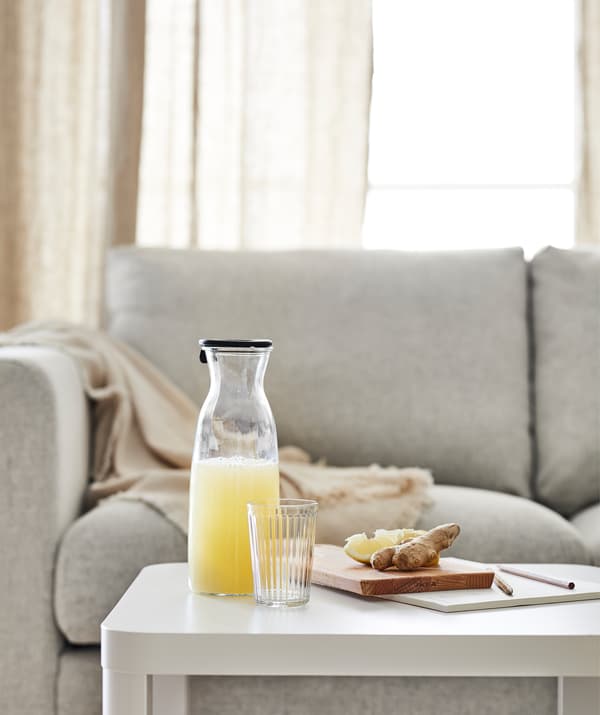 Una caraffa KARAFF con una bevanda alla frutta e un tagliere con fette di limone e zenzero appoggiati su un tavolino TINGBY, davanti a un divano.