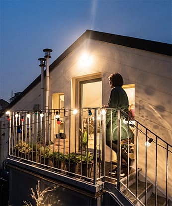 Una casa vista da fuori, con la facciata con tetto a punta e il balconcino. Dentro le luci sono accese e si sente atmosfera di casa. La porta è aperta. Una persona sta per entrare.
