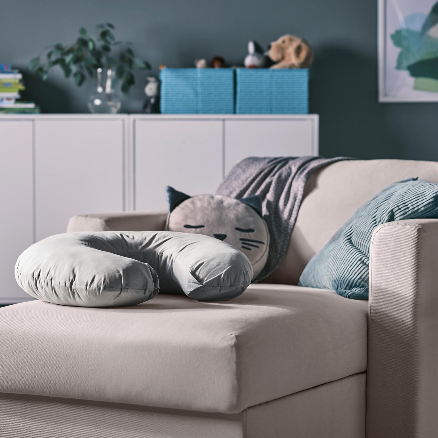 Una chaise longue beige con un cojín de lactancia gris y varios cojines, además de armarios blancos con cajas y un jarrón encima.
