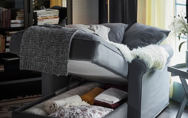 Una chaise longue grigia GRÖNLID con il contenitore alzato. All'interno coperte, cuscini e altri accessori. Nella stanza sul lato sinistro si trova una vetrina con ante. Sulla destra si intravede un tavolino con sopra un vaso. 