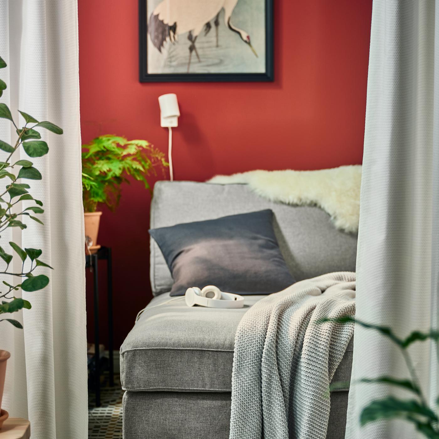 Una chaise-longue KIVIK grigia con un plaid INGABRITTA grigio chiaro e un cuscino antracite, più una lampada da parete NYMÅNE bianca.