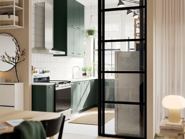 Una cocina abierta con frentes de cocina HAVSTORP en verde oscuro y gabinetes de pared hasta el techo junto a una sala luminosa.