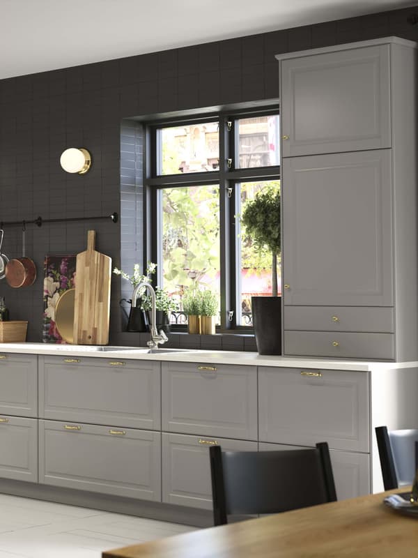 Una cocina BODBYN gris organizada con una tabla de cortar en la encimera y una gran ventana sobre el fregadero.
