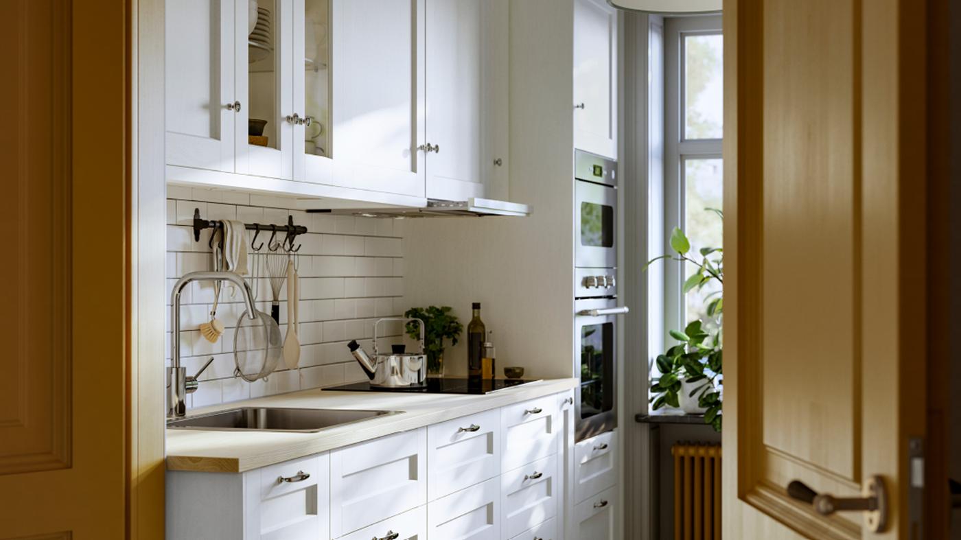 Una cocina con cajones y armarios de pared con frentes ENKÖPING blancos y un riel HULTARP niquelado con ganchos en la pared.