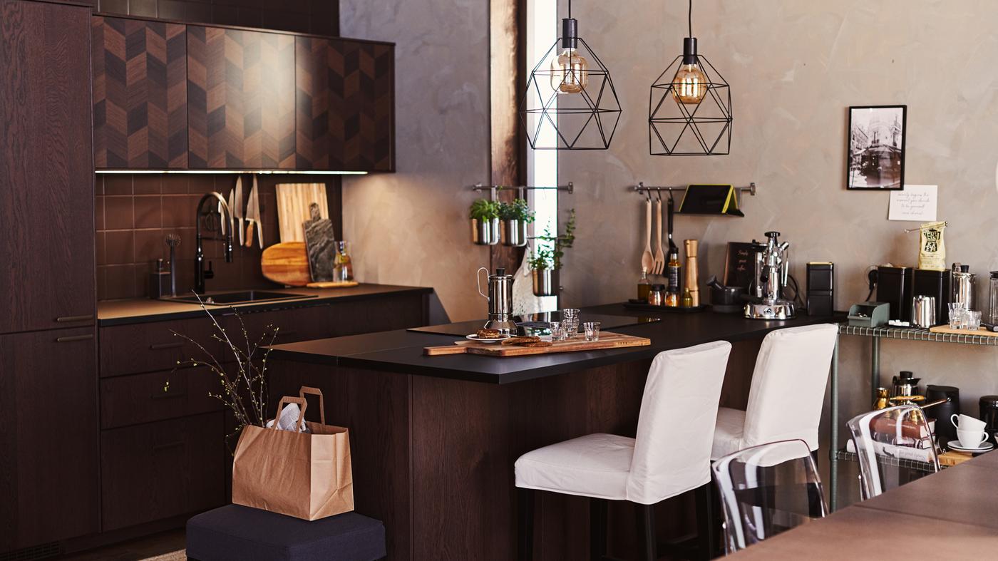 Una cocina HASSLARP/SINARP en marrón oscuro con isla. Hay taburetes altos blancos en torno a la isla y dos lámparas BRUNSTA colgadas encima.