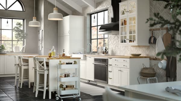 Una cocina SEKTION con puertas BODBYN y puertas de cristal en hueso blanco, un carrito RÅSKOG, lámparas BUNKEFLO de techo en beige/abedul.