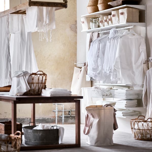 Una combinazione di guardaroba BOAXEL bianca con vestiti appesi a grucce bianche e cestini sulla parte superiore, più un tavolo trattato con mordente marrone - IKEA.