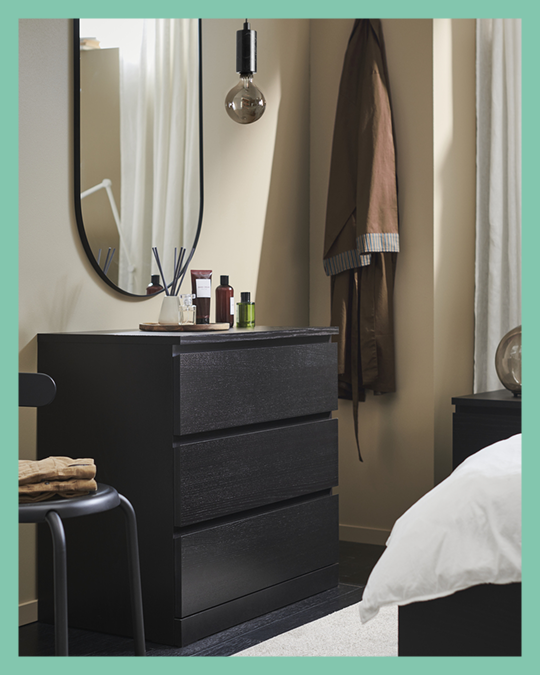 Una cómoda IKEA negra de 3 cajones frente a una cama. En la pared de la cómoda un espejo ovalado.