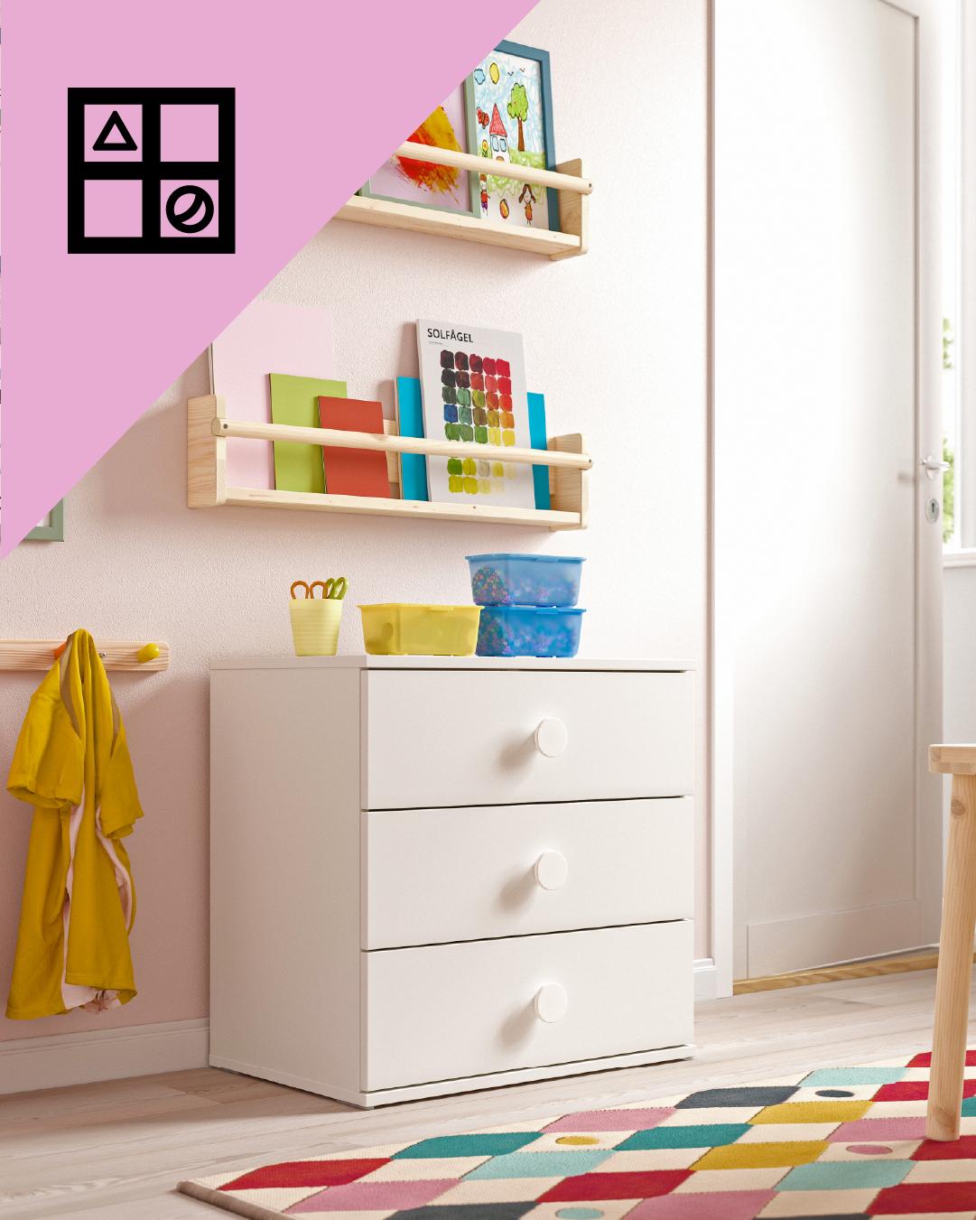 Una cómoda infantil blanca con tres cajones y sobre ella cajas organizadoras frente a una alfombra colorida. En la esquina superior de la imagen un triángulo rosa con figuras geométricas.