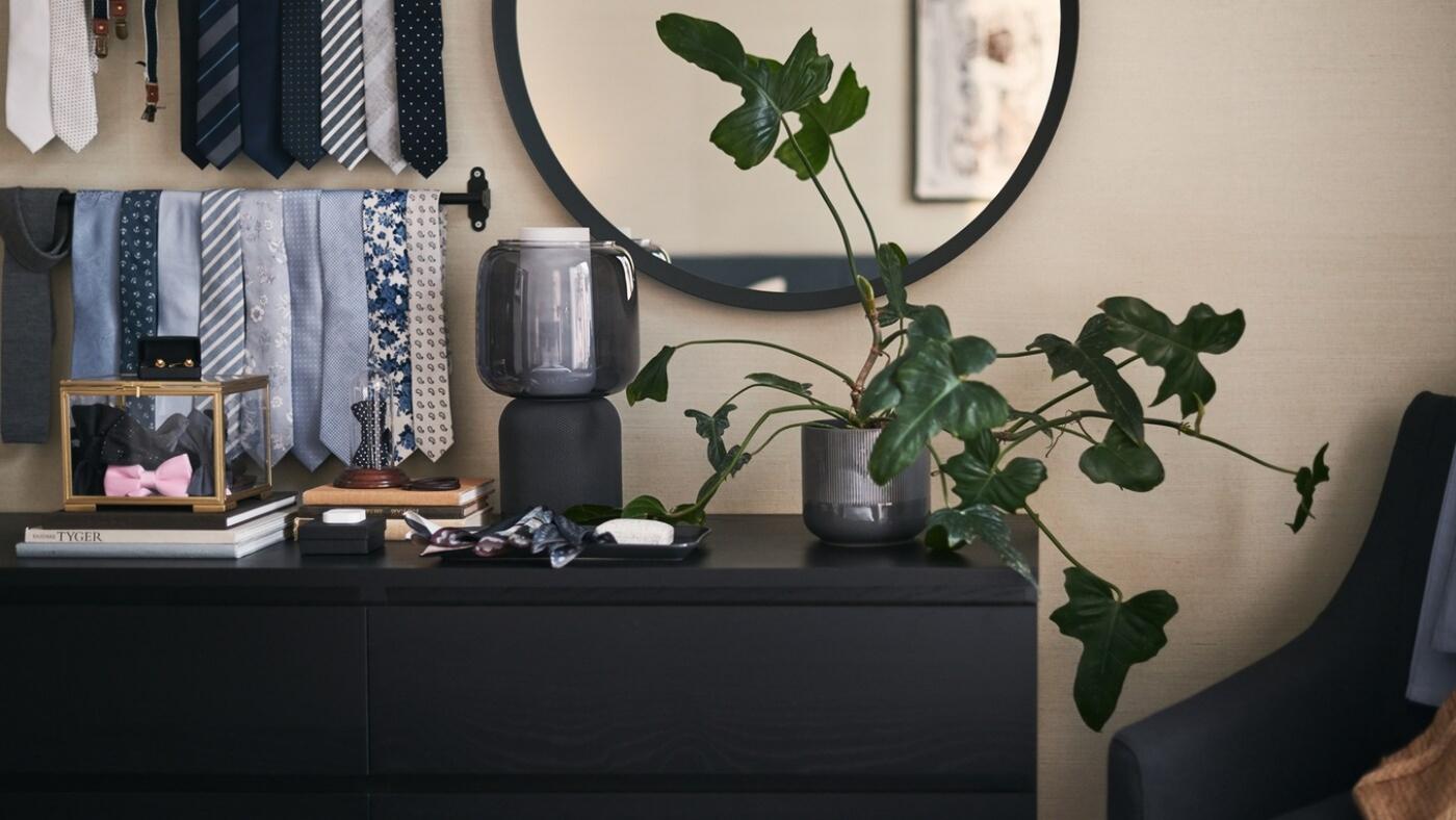 Una cómoda MALM en negro-marrón delante de un gran espejo redondo con una planta frondosa y un altavoz wifi SYMFONISK encima.