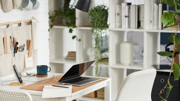 Una computadora portátil sobre una mesa blanca con una silla blanca y otra beige, junto a una unidad de estantería blanca con libros, plantas y almacenamiento.