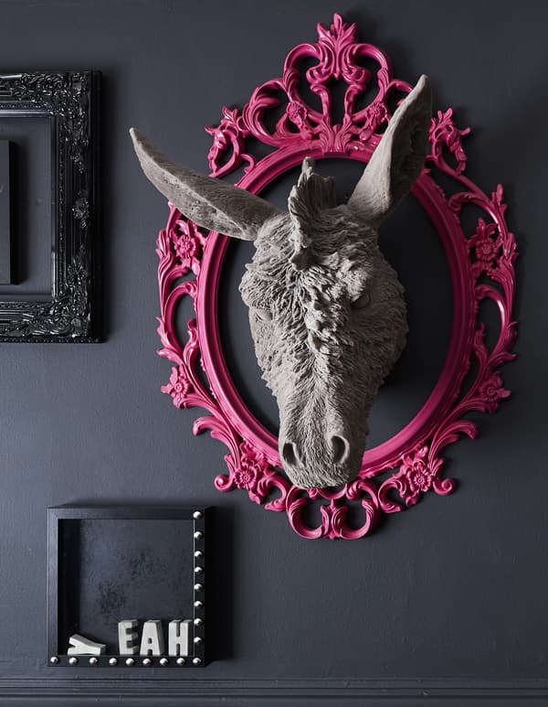 Una cornice rosa fluo su una parete nera con dentro la testa di un asino scolpita.