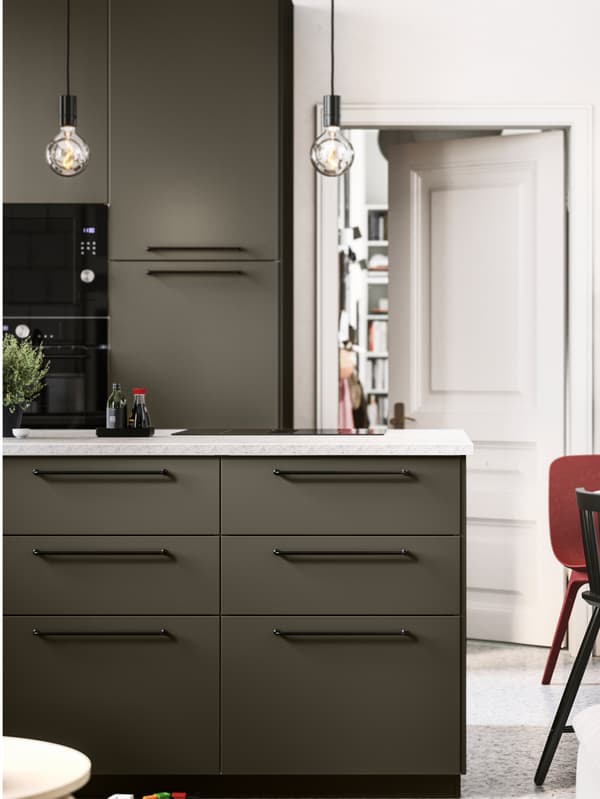 Una cucina a vista con frontali HAVSTORP marrone-beige, maniglie ed elettrodomestici neri e piano di lavoro effetto pietra calcarea.