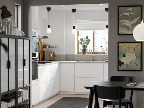 Una cucina con ante e frontali VOXTORP bianco opaco lisci, tre lampade a sospensione nere in fila e un’area sala da pranzo in primo piano - IKEA