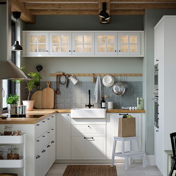 Una cucina con frontali per cassetti STENSUND e ante a vetro, piani di lavoro effetto rovere e un lavello con frontale visibile bianco.