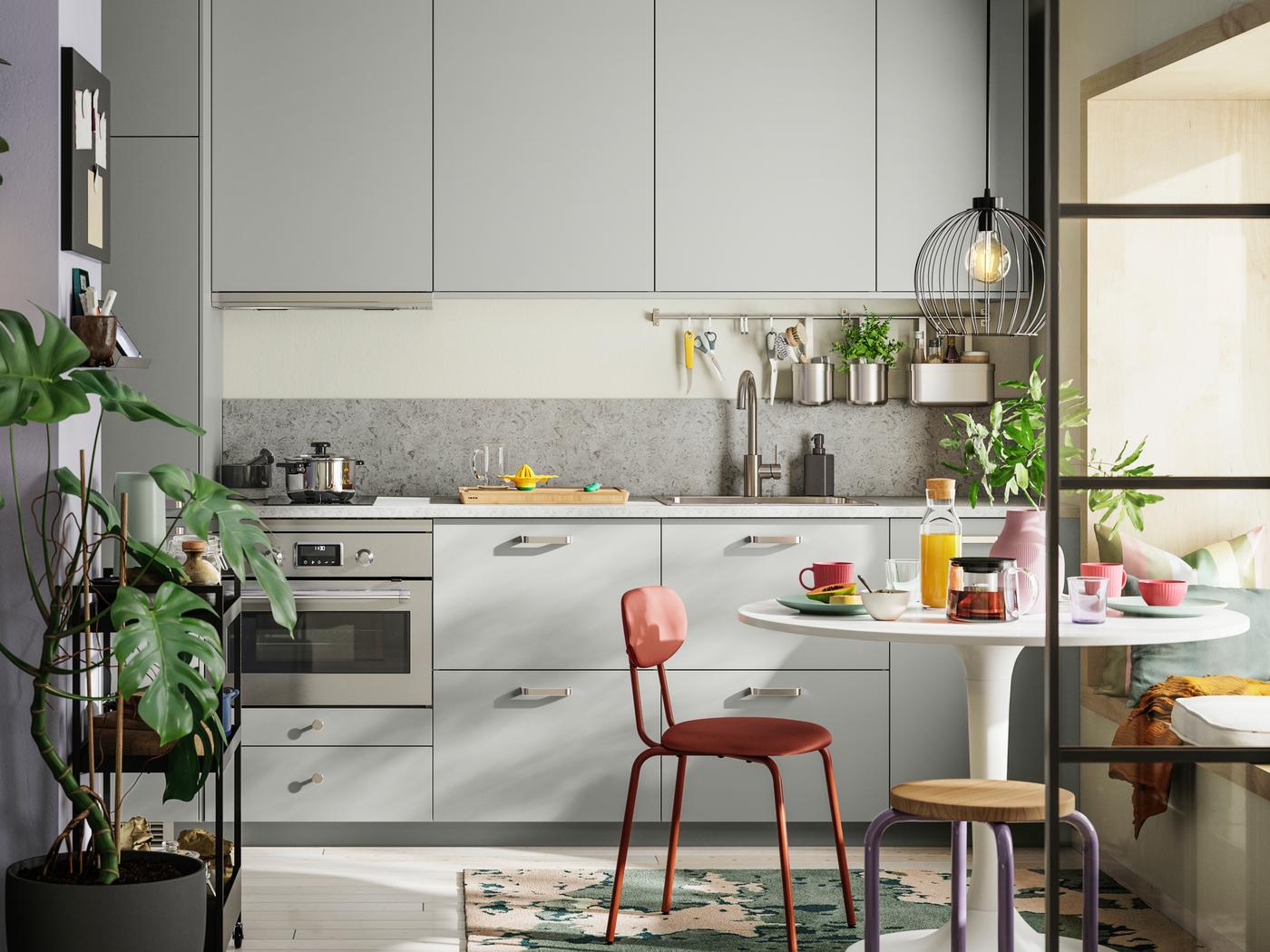 Una cucina lineare con frontali HAVSTORP grigio chiaro arredata da un tavolo DOCKSTA bianco, una sedia ÖSTANÖ rosso-marrone e uno sgabello DOMSTEN.