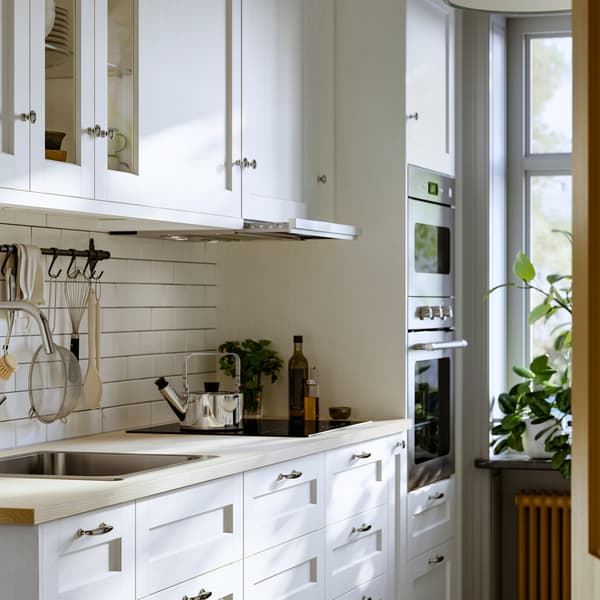 Una cucina luminosa con le piastrelle bianche e una finestra include vari mobili con frontali ENKÖPING bianchi ed elettrodomestici in acciaio inossidabile.