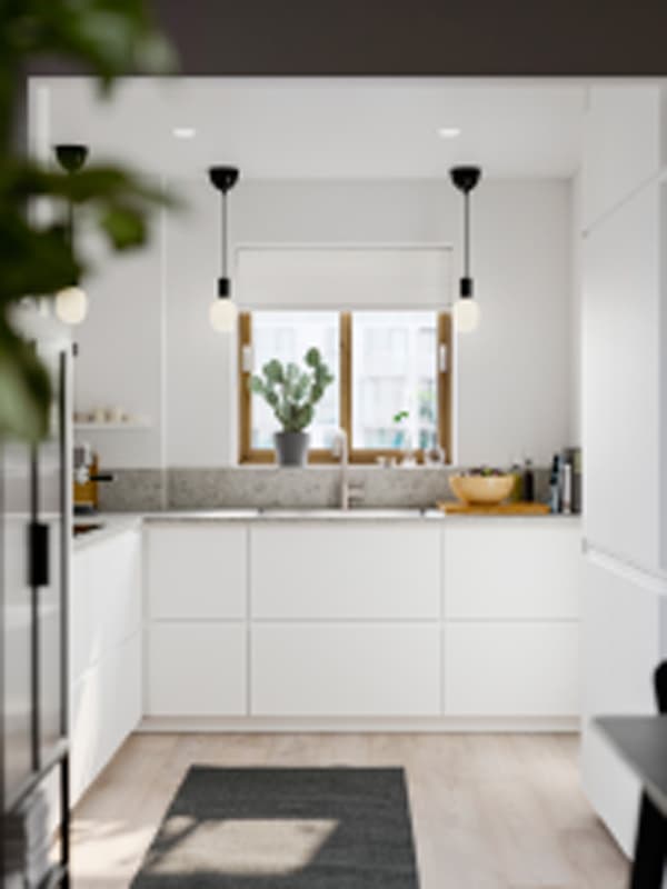 Una cucina piccola e luminosa con frontali VOXTORP bianco opaco, pareti bianche, un tappeto grigio e lampade a sospensione nere.
