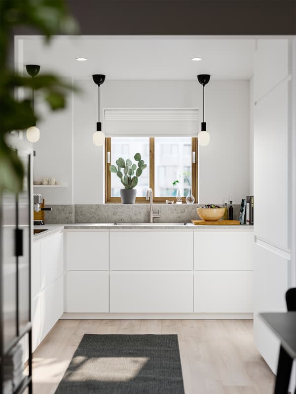 Una cucina piccola e luminosa con frontali VOXTORP bianco opaco, pareti bianche, un tappeto grigio e lampade a sospensione nere.