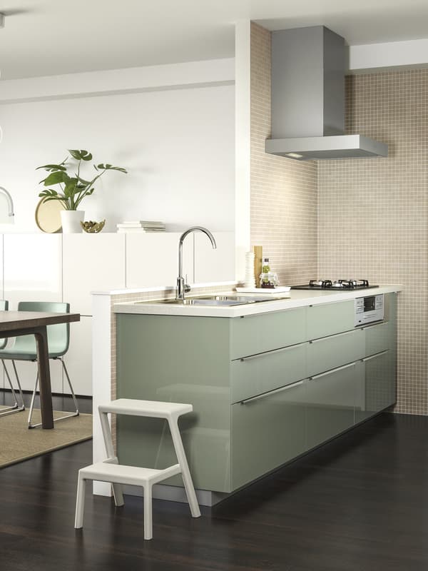 Una cucina sui toni del verde con cappa in alluminio appoggiata ad un muretto. Si intravede un soggiorno sui toni del bianco ben illuminato.