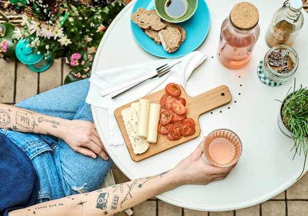 Una dona amb els braços tatuats subjecta un got i seu en una taula rodona de color crema amb una safata amb formatge i tomàquets.