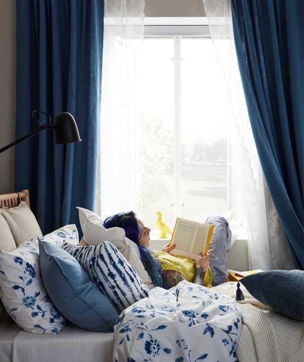 Una donna appoggiata a un mucchio di morbidi cuscini legge un libro a letto con i piedi sul davanzale della finestra.