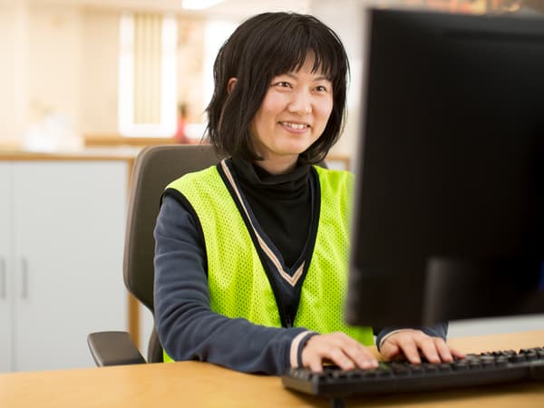 Una donna che indossa un gilet di sicurezza lavora al computer. Sorride.